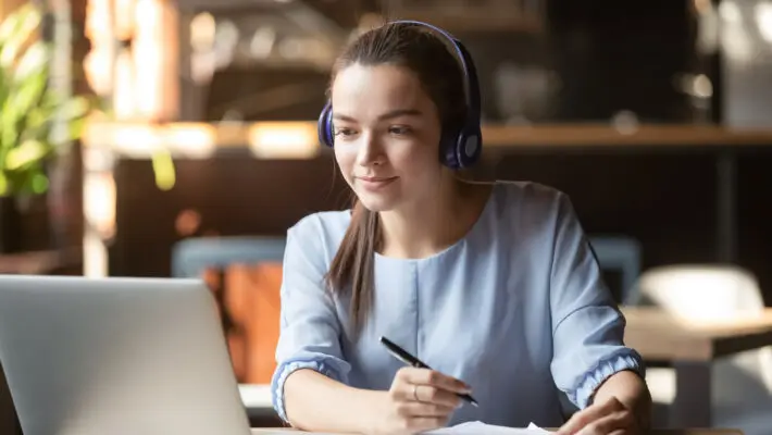 Focused,Woman,Wearing,Headphones,Using,Laptop,In,Cafe,,Writing,Notes,