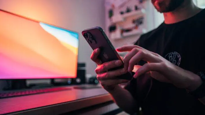 Man holding an iPhone in front of his desk, with a monitor in the background.